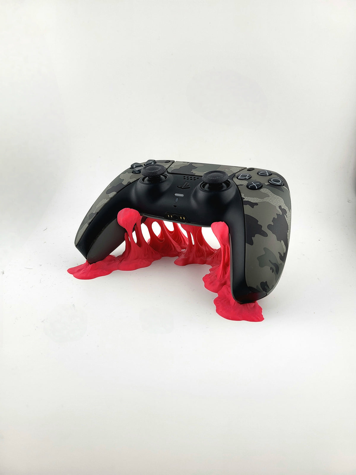 Front three-quarter view of a red 3D printed slime stand supporting a camouflage PS5 DualSense controller above rising tendrils.