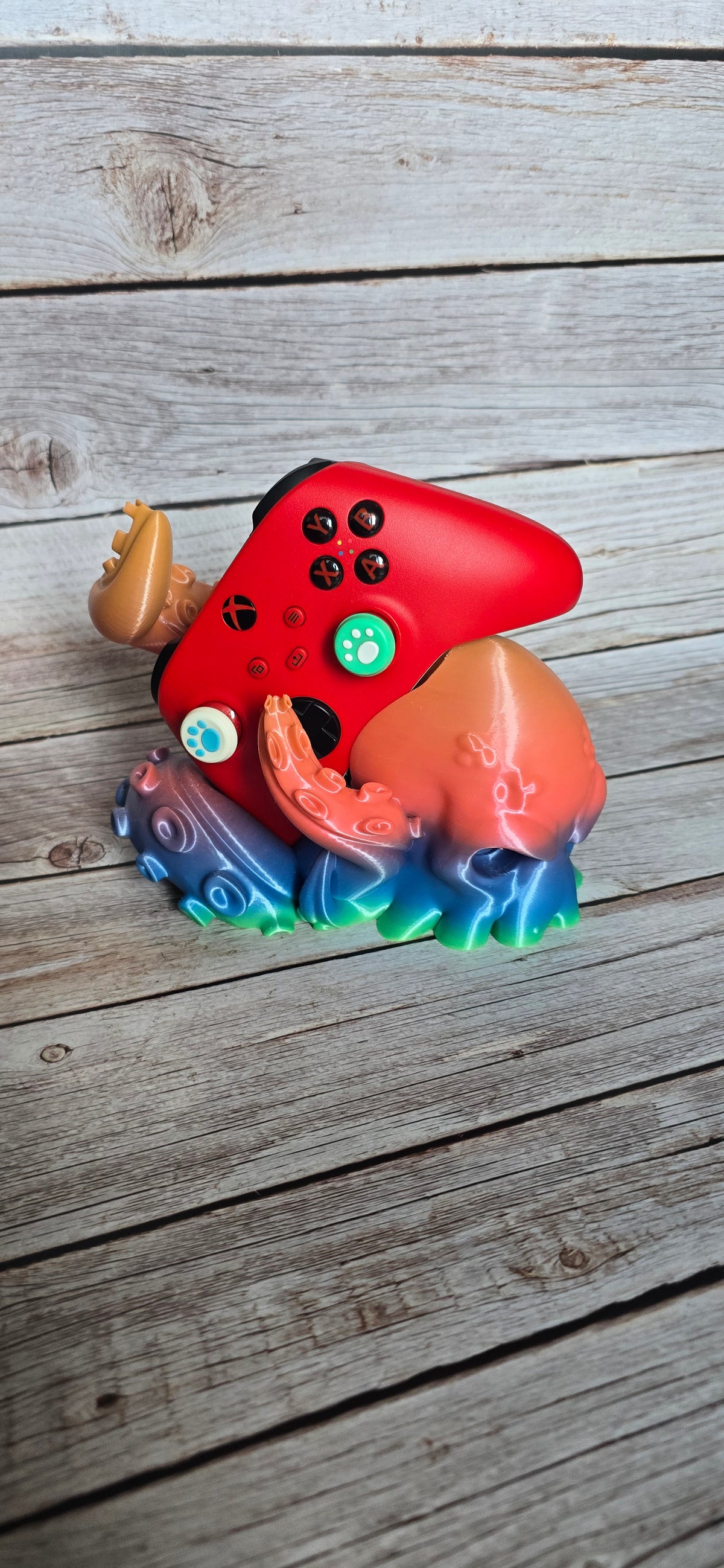Front three-quarter view of a colourful Kraken controller stand holding a red Xbox-style controller between curled tentacles.