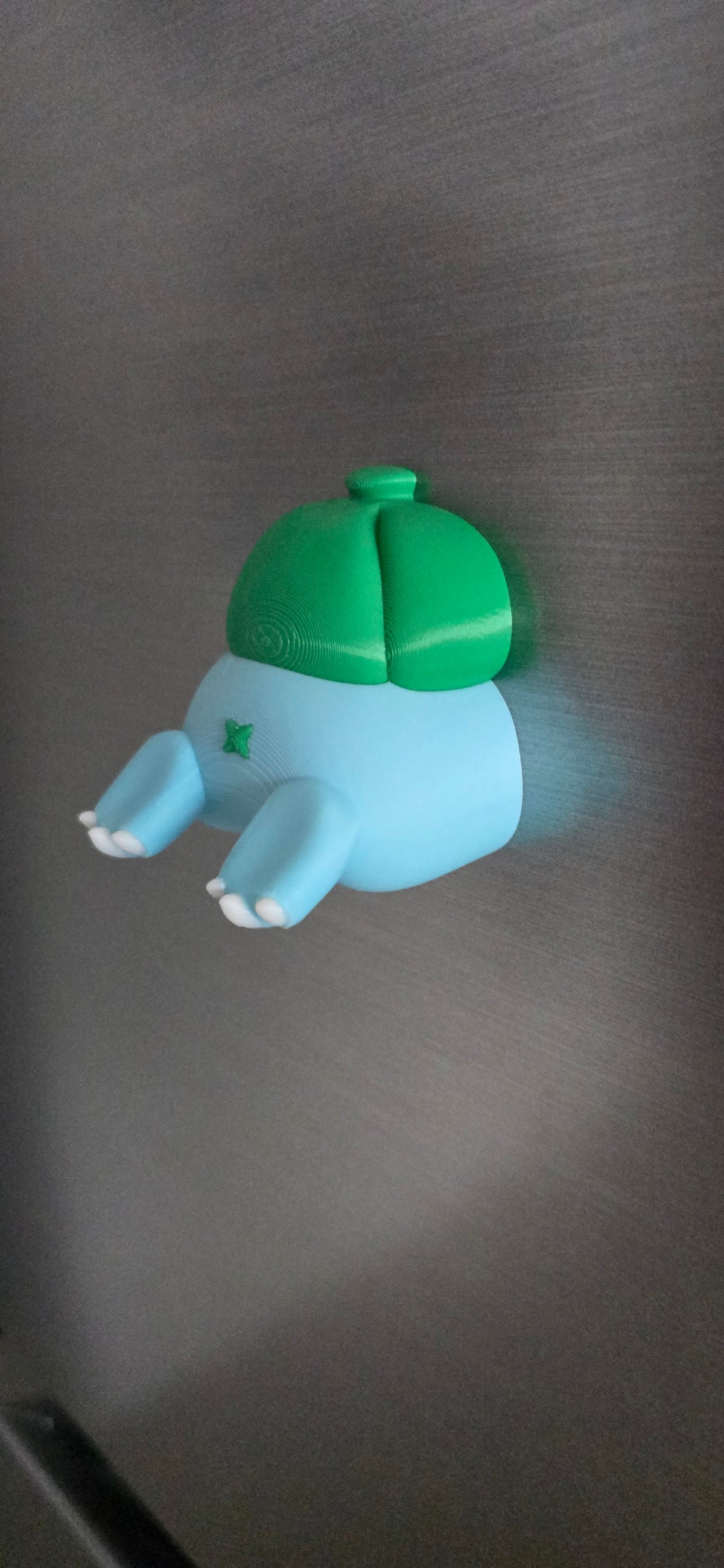 Front view of a 3D printed blue creature fridge magnet with tiny white claws and a green bulb on its back, creating a funny burrowing illusion.
