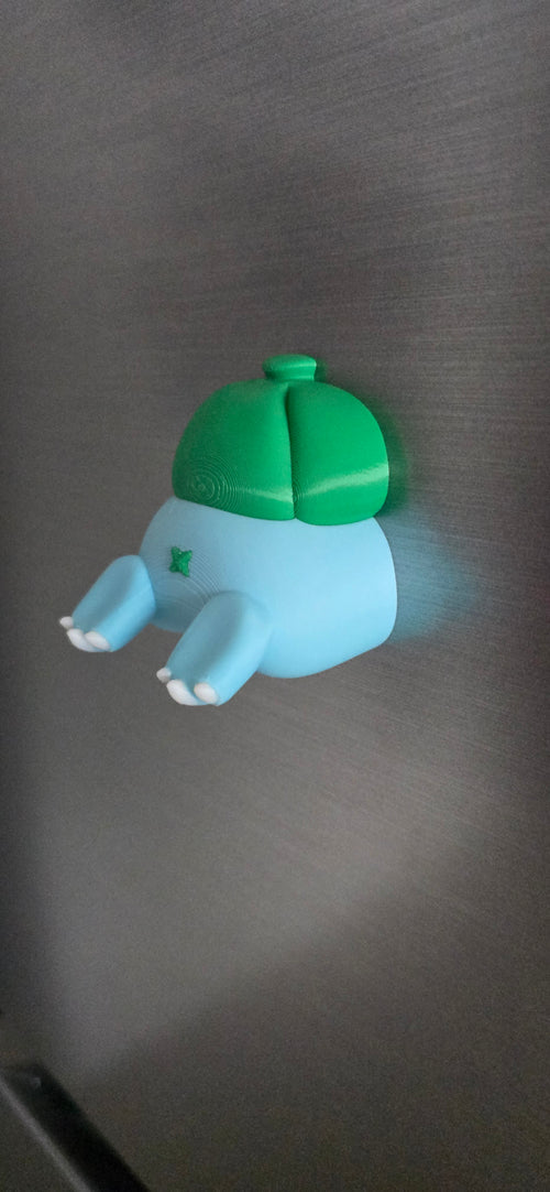 Front view of a 3D printed blue creature fridge magnet with tiny white claws and a green bulb on its back, creating a funny burrowing illusion.