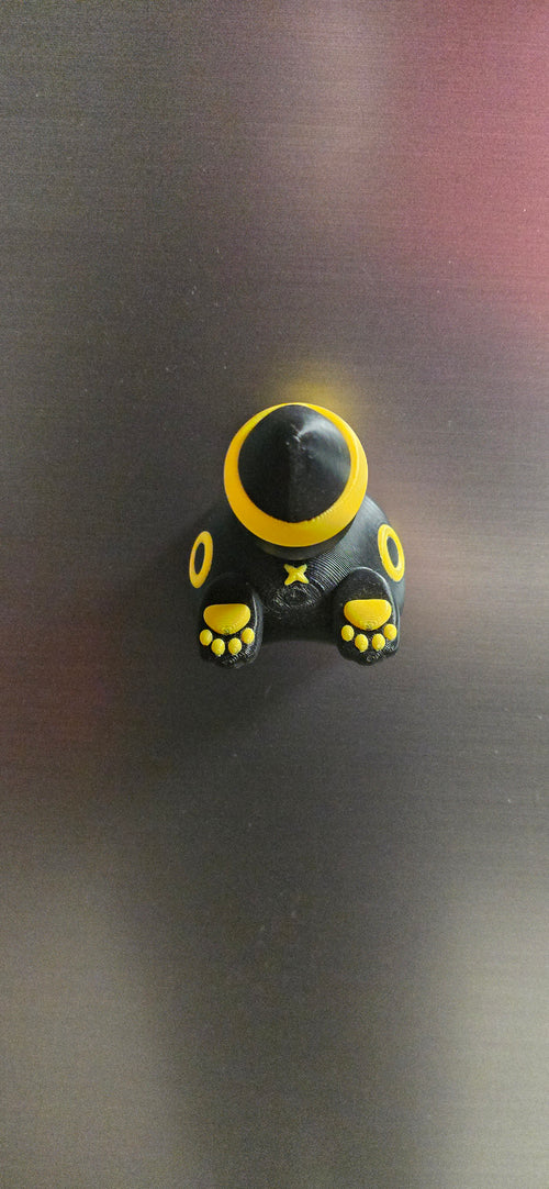 Front-angled view of the Nocturne Burrow Familiar magnet showing its yellow body rings, curled tail and two small black paws with yellow pads.
